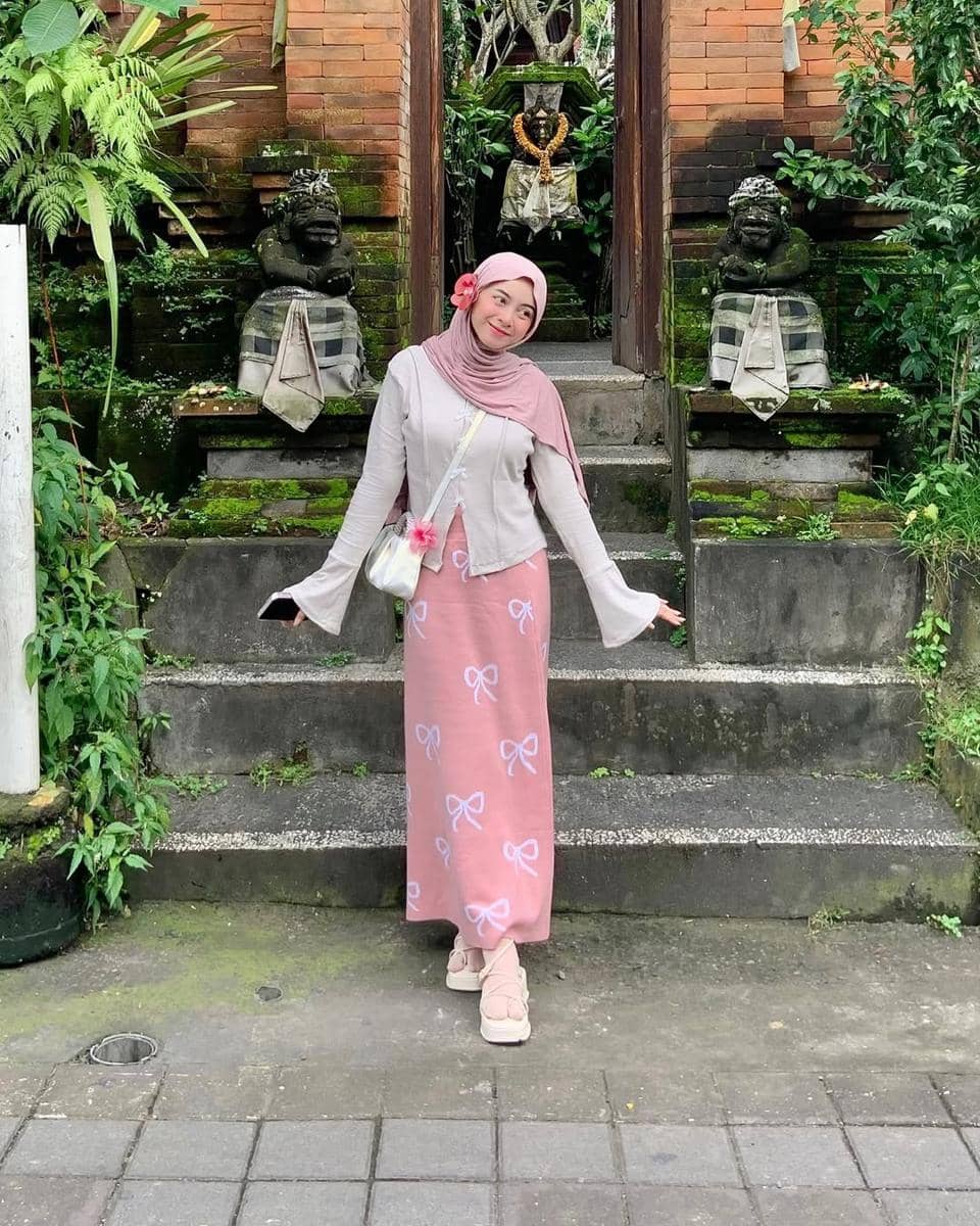 Seorang wanita berhijab berpose ceria di depan gerbang batu bergaya Bali dengan patung tradisional dan tanaman hijau di sekitarnya.