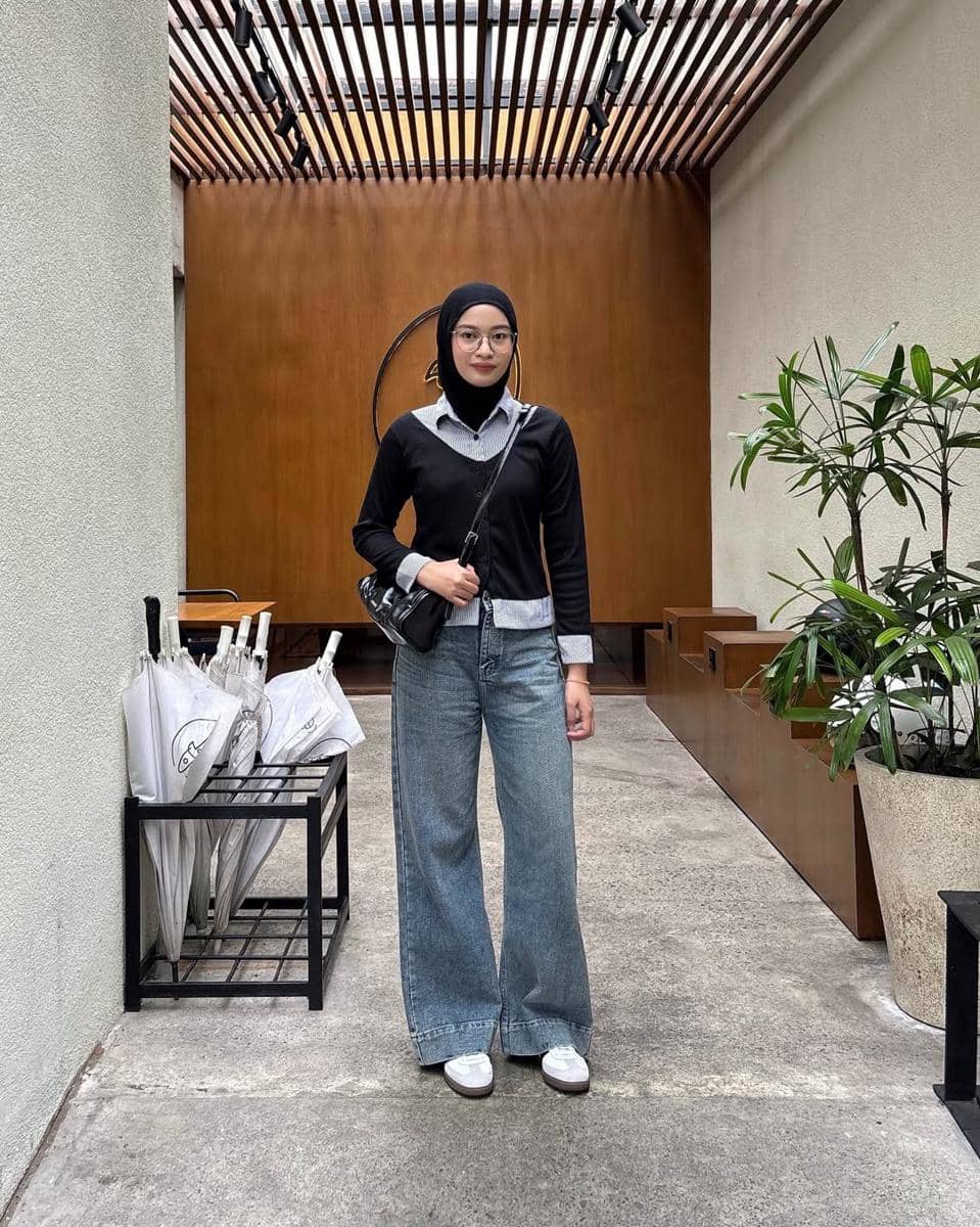 Seorang remaja berhijab mengenakan kemeja berlapis sweater hitam, celana jeans longgar, dan sepatu putih berdiri di lorong bergaya minimalis.