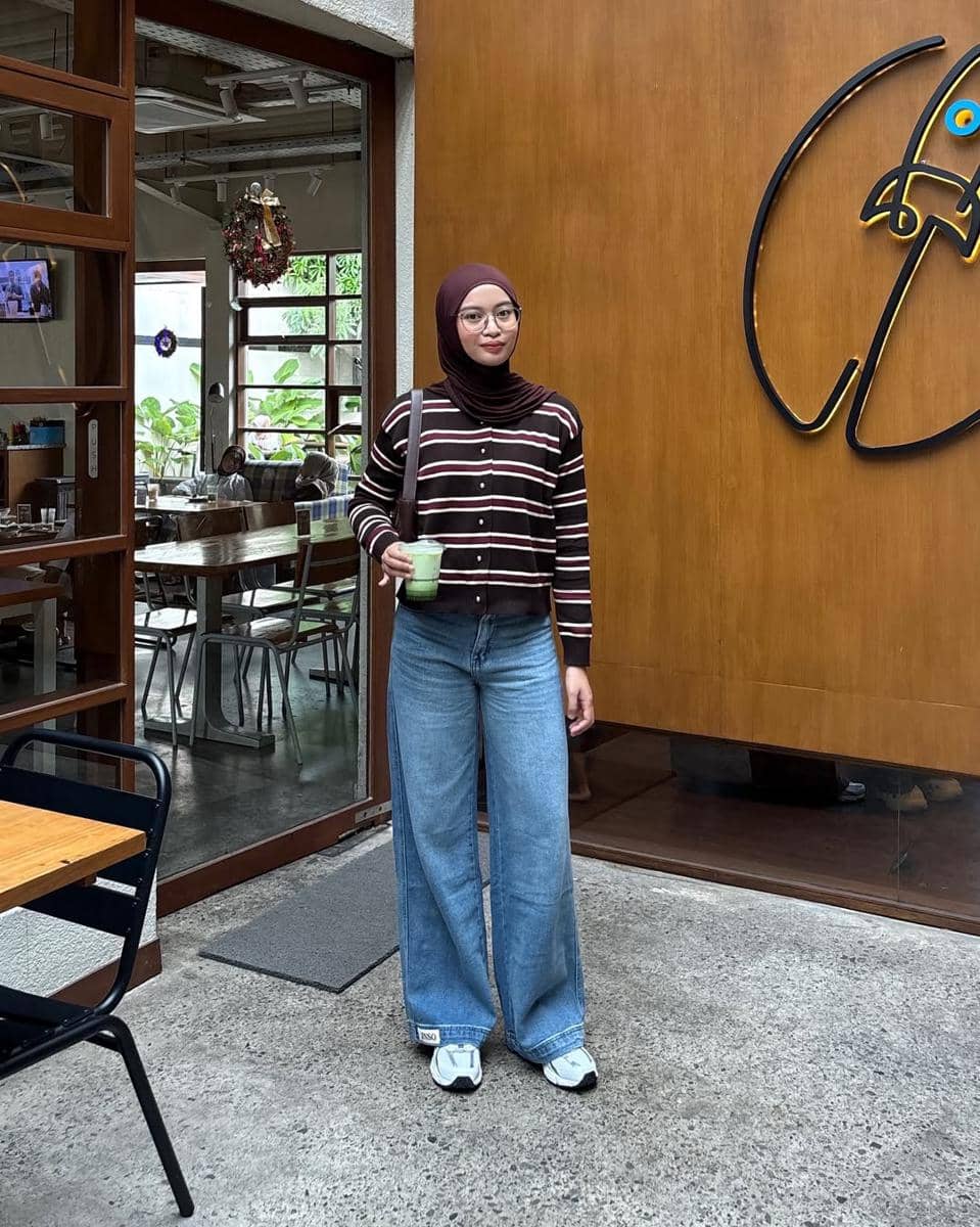 Seorang remaja berhijab mengenakan kardigan garis-garis cokelat dan celana jeans longgar sambil memegang minuman di depan kafe.