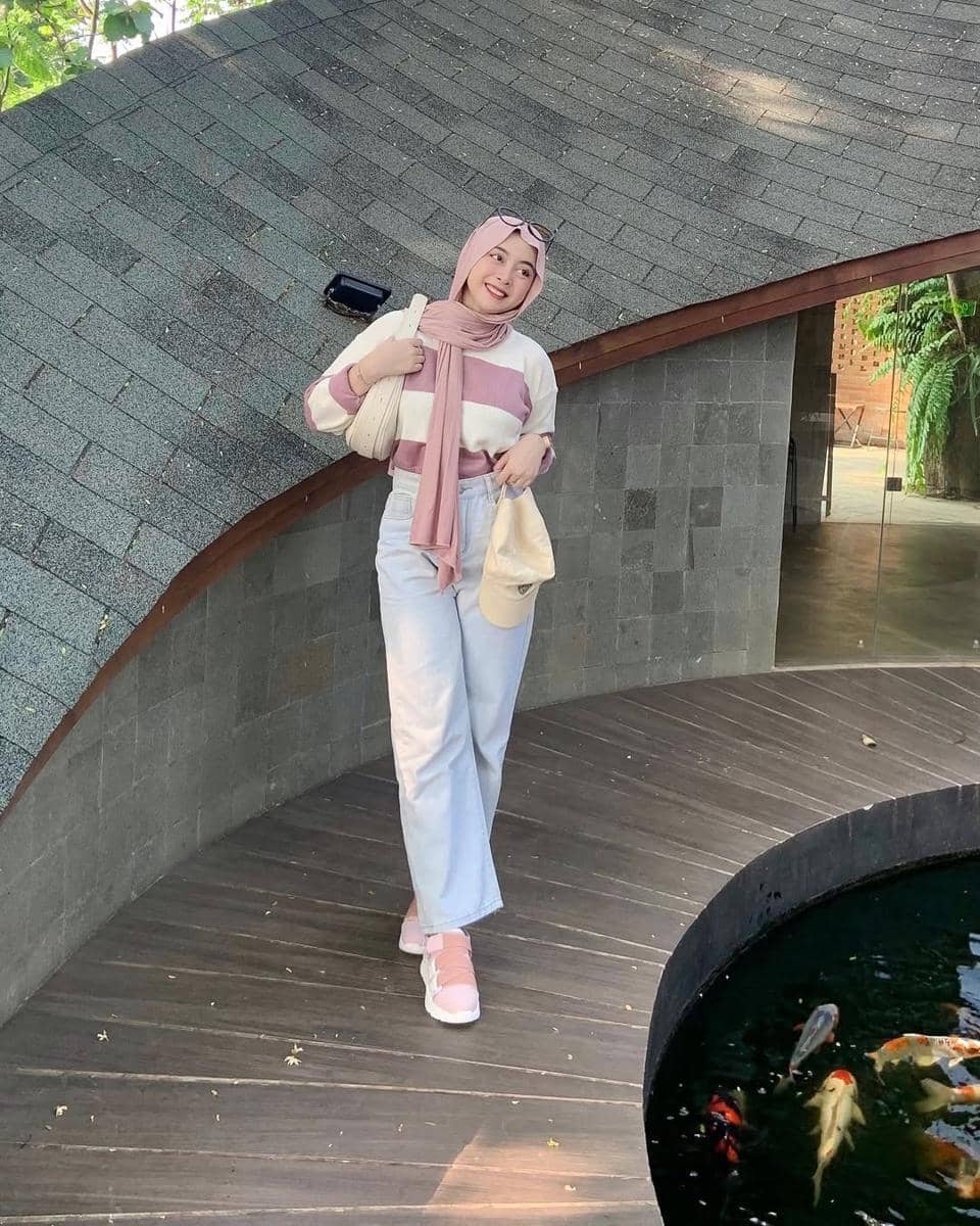 Seorang wanita berhijab mengenakan sweater bergaris merah muda dan putih, celana panjang terang, serta sepatu merah muda di dekat kolam ikan koi.