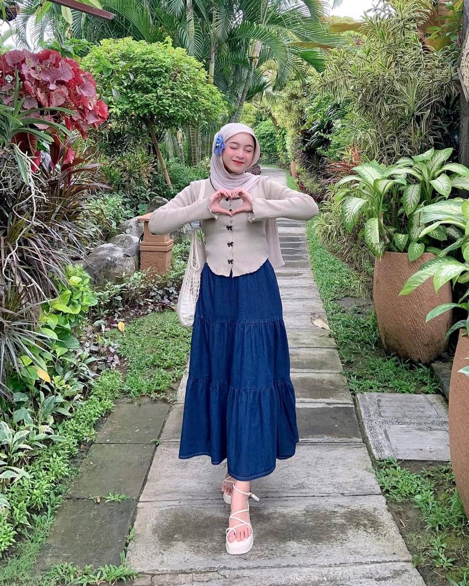 Seorang wanita berhijab berpose membentuk hati dengan tangan di taman hijau, mengenakan rok denim panjang dan atasan krem.