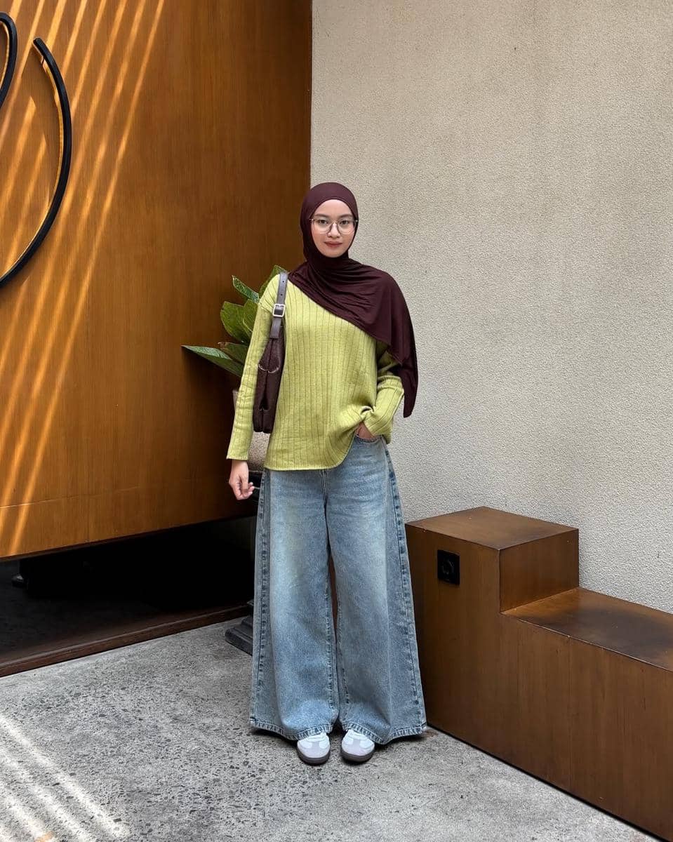 Seorang wanita berhijab cokelat mengenakan sweater hijau muda dan celana jeans longgar berdiri di area outdoor dengan dinding kayu dan beton.