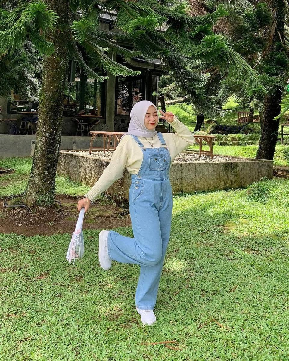 Seorang wanita berhijab berpose ceria di taman hijau mengenakan overall denim biru muda, sweater krem, dan sepatu putih.
