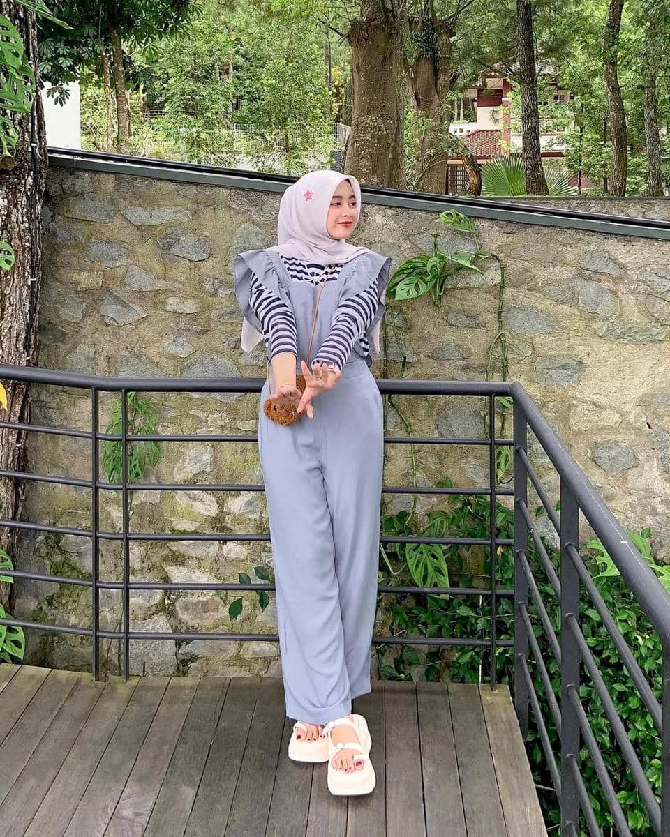 Seorang wanita berhijab berpose santai di balkon kayu dengan latar tembok batu dan pepohonan hijau, mengenakan outfit kasual berwarna abu-abu.