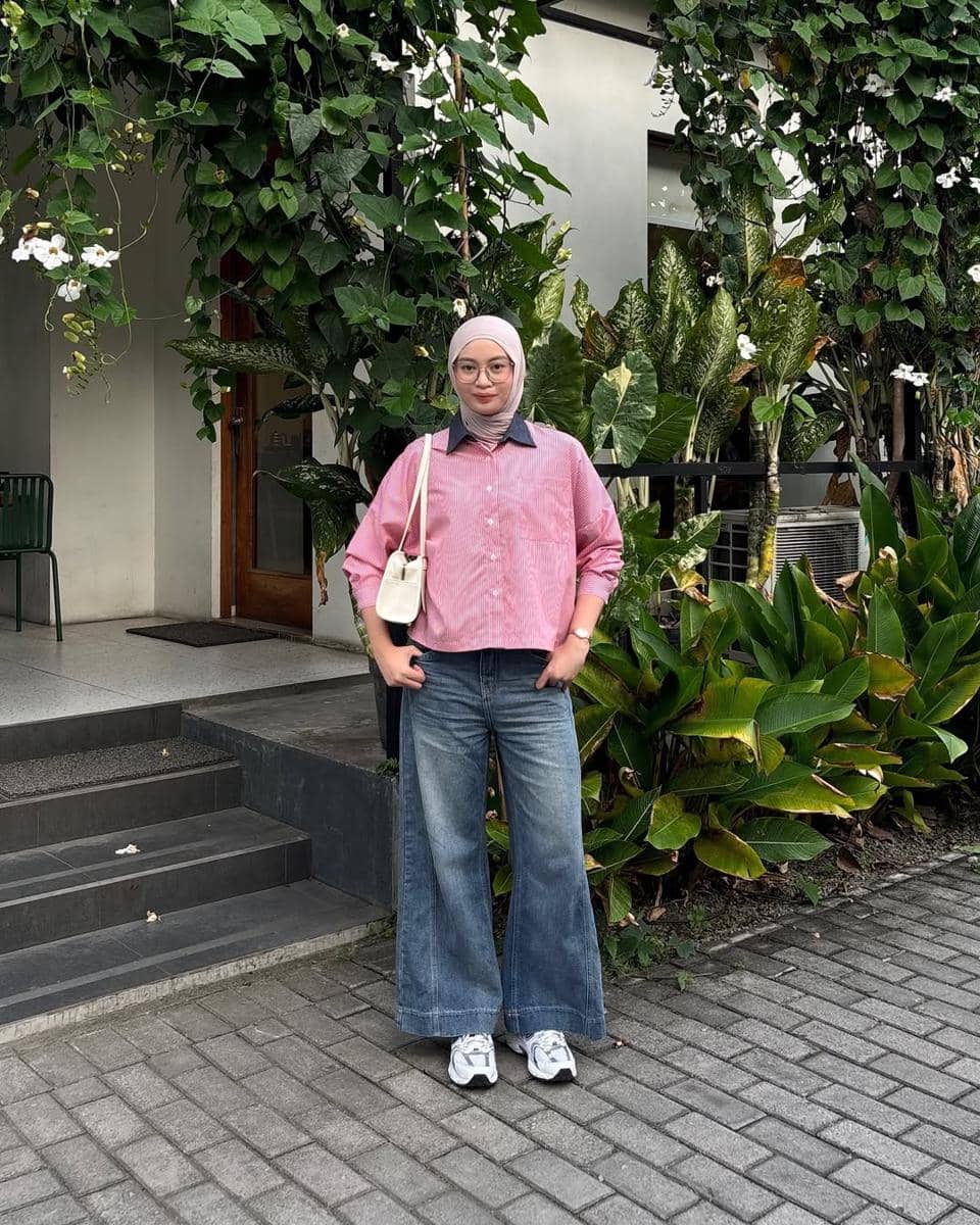 Seorang perempuan berhijab mengenakan kemeja merah muda, celana jeans lebar, dan sepatu putih berpose di depan bangunan dengan tanaman hijau.