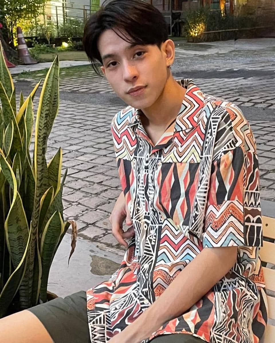 Seorang pria mengenakan kemeja bermotif warna-warni dengan celana pendek hijau duduk di bangku dekat tanaman di area jalan berbatu.