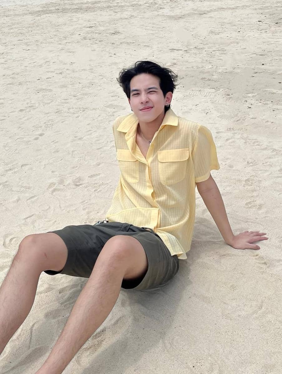 Seorang pria mengenakan kemeja kuning dan celana pendek duduk santai di atas pasir pantai dengan ekspresi tersenyum.