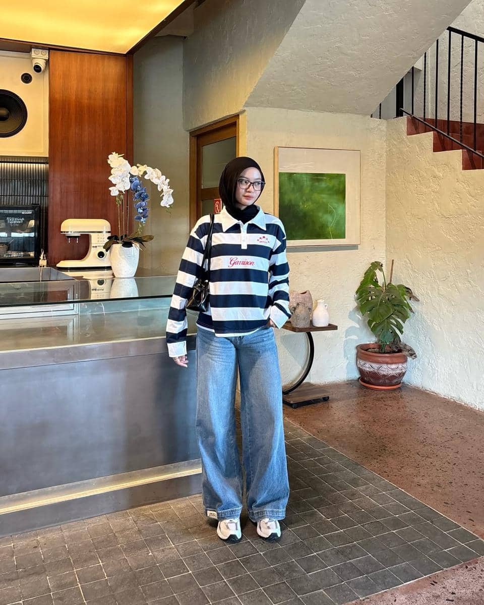 Seorang remaja berhijab mengenakan kaus garis-garis biru putih, celana jeans longgar, dan sepatu sneakers berdiri di dalam kafe modern.