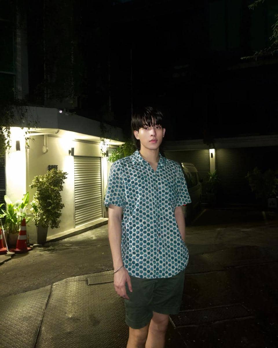 Seorang pria mengenakan kemeja motif polkadot biru dan celana pendek hijau berdiri di area luar gedung pada malam hari.