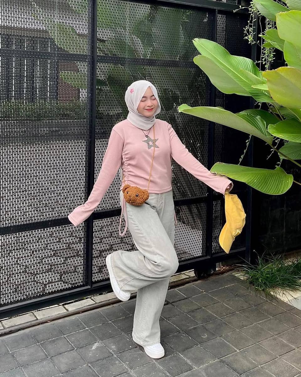 Seorang perempuan berhijab bergaya kasual dengan atasan pink dan celana abu-abu berpose ceria di depan pagar hitam dan tanaman hijau.