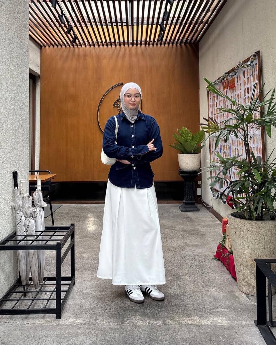 Seorang remaja berhijab bergaya kasual mengenakan kemeja denim biru tua, rok putih panjang, dan sepatu sneakers putih di area indoor modern.
