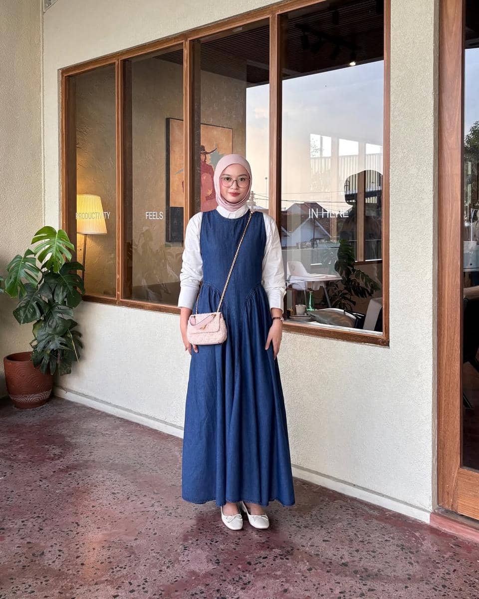 Seorang wanita berhijab mengenakan long dress denim biru dengan kemeja putih dan tas kecil berwarna pink di depan kafe modern.