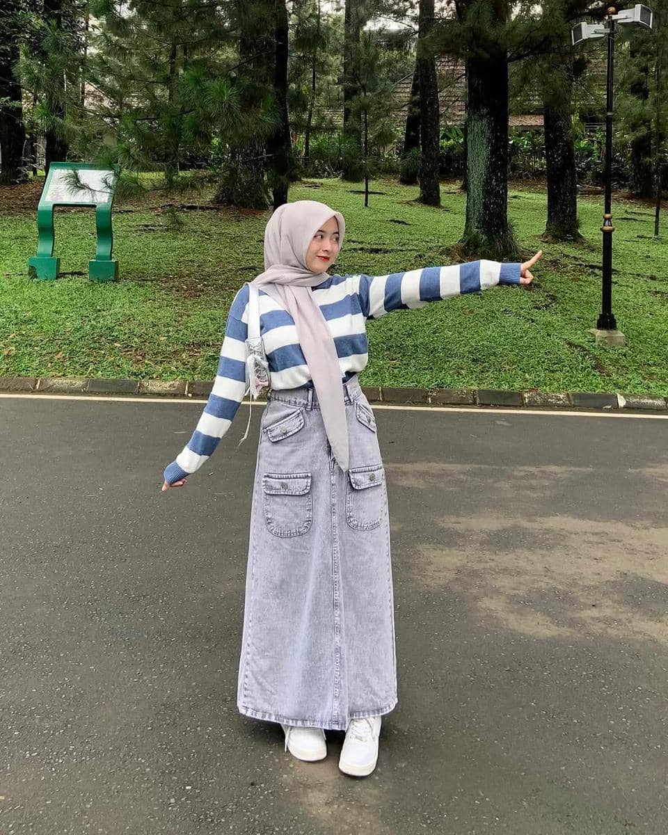 Seorang wanita berhijab bergaya kasual dengan sweater garis biru putih dan rok denim panjang berwarna abu-abu di area taman hijau.