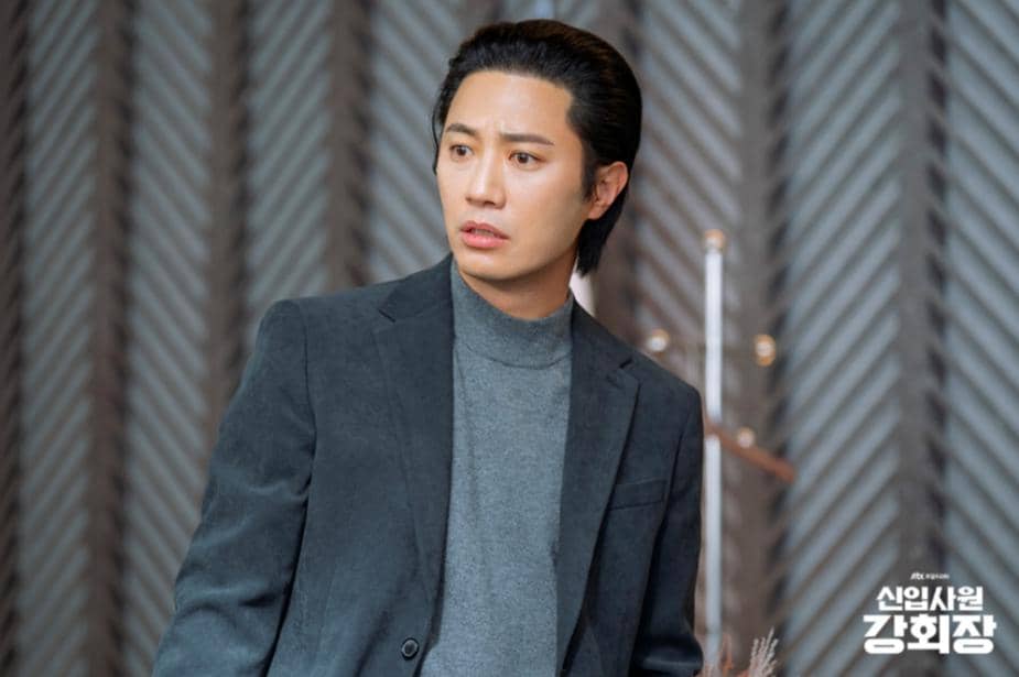 Seorang pria mengenakan jas abu-abu gelap dan turtleneck abu-abu berdiri di dalam ruangan dengan latar dinding berpola diagonal.