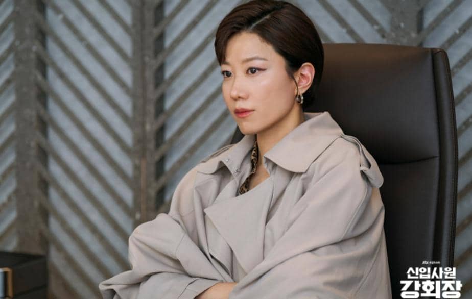 Seorang wanita berambut pendek mengenakan mantel abu-abu duduk di kursi kantor dalam cuplikan drama Reborn Rookie.