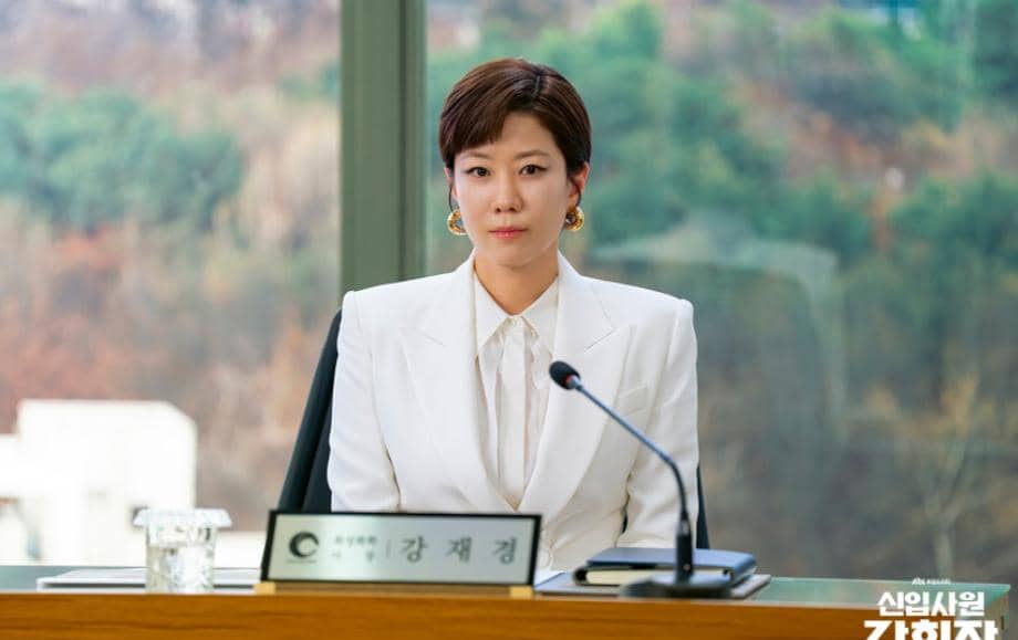 Seorang wanita mengenakan setelan putih duduk di meja konferensi dengan mikrofon di depannya dalam cuplikan drama Reborn Rookie.