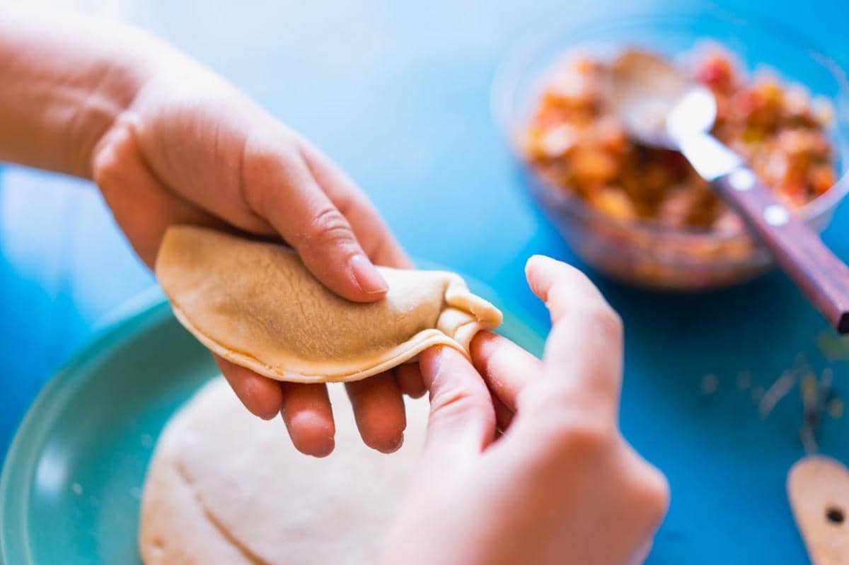 Tangan seseorang sedang melipat adonan empanada di atas piring dengan isian sayuran di mangkuk kaca di latar belakang.