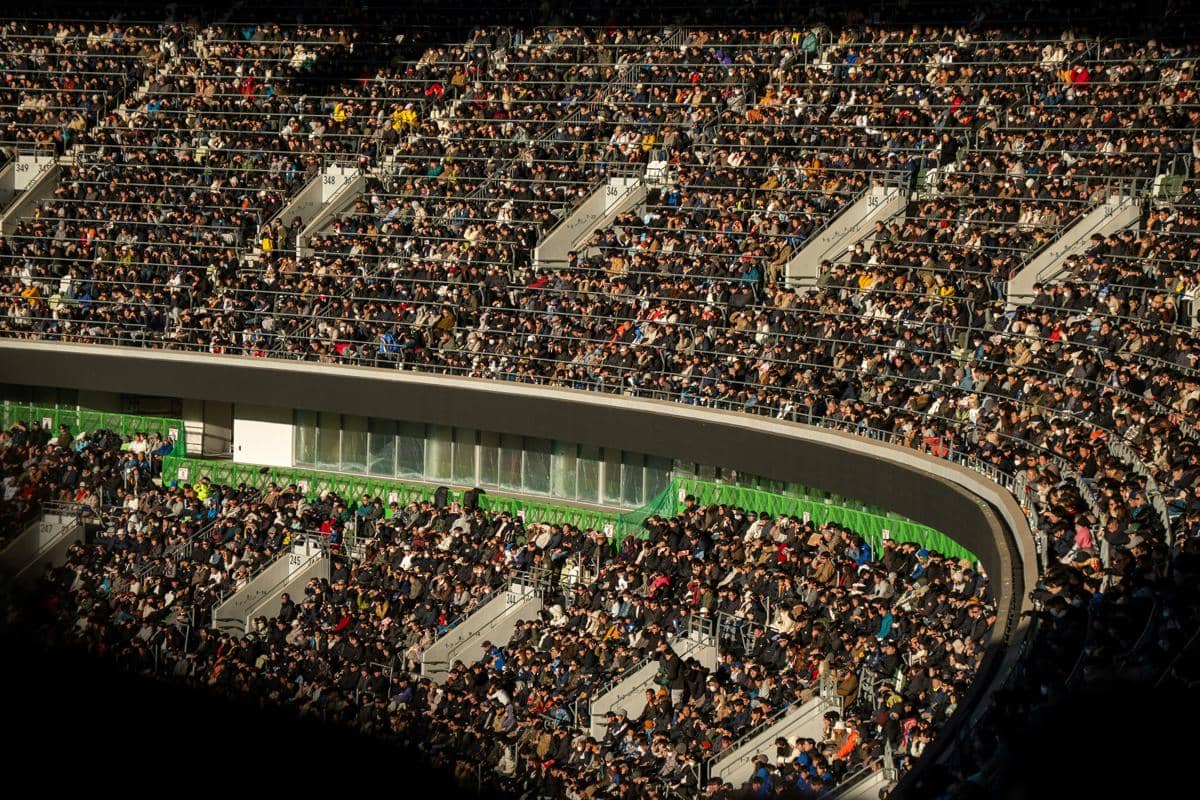 Pemandangan dari sudut atas tribun stadion megah bertingkat dua yang dipenuhi oleh ribuan penonton sepak bola yang duduk rapat di kursi mereka saat siang hari.