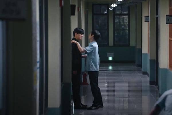 Dua siswa berdiri di lorong sekolah dengan pencahayaan redup, salah satunya memegang bahu yang lain dalam suasana tegang.