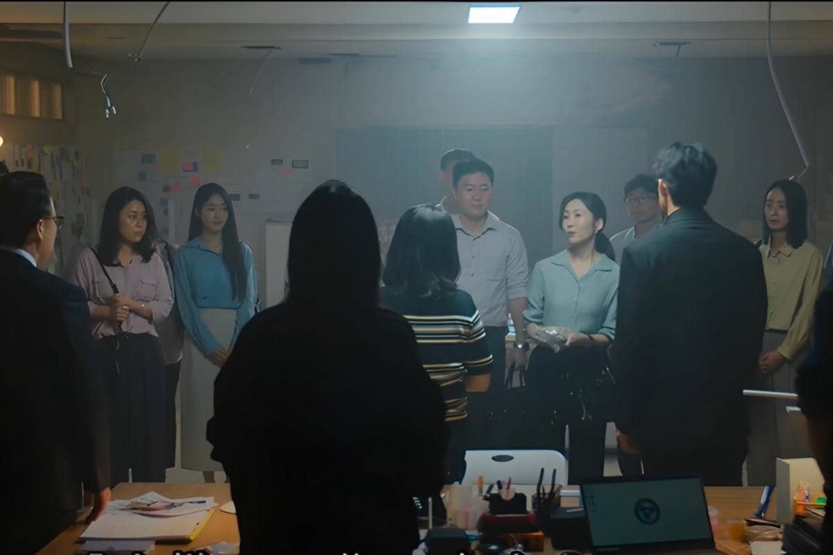 Beberapa orang berdiri melingkar di dalam ruangan kantor dengan pencahayaan redup dalam adegan drama Teach You a Lesson.
