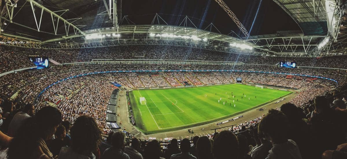 Pemandangan sudut lebar dari tribun penonton yang penuh sesak di dalam stadion sepak bola megah modern dengan lampu sorot cerah pada malam hari.