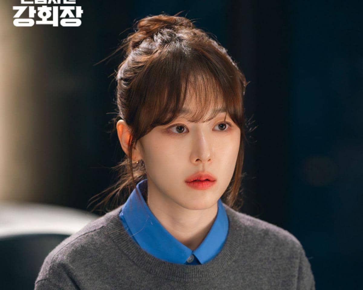 Seorang wanita berambut cokelat dengan poni mengenakan kemeja biru dan sweater abu-abu tampak serius dalam cuplikan drama Reborn Rookie.