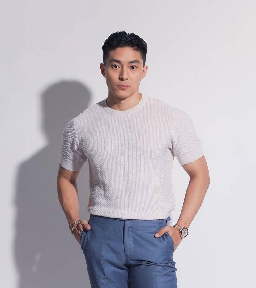 Seorang pria berdiri dengan tangan di saku, mengenakan kaus putih dan celana panjang biru di latar belakang putih polos.