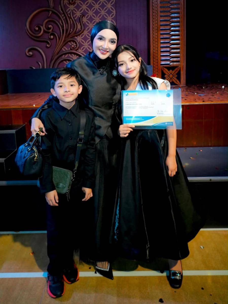 Seorang perempuan berbusana hitam berpose bersama dua anak yang juga mengenakan pakaian hitam, salah satunya memegang ijazah kelulusan SD.