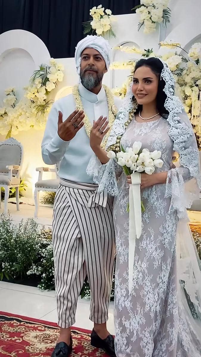 Pasangan pengantin berdiri di pelaminan dengan dekorasi bunga putih, mempelai pria mengenakan pakaian adat dan mempelai wanita mengenakan gaun renda sambil memegang buket bunga.