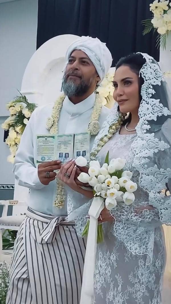Sepasang pengantin mengenakan busana adat berwarna putih berpose sambil memegang buku nikah dan buket bunga di pelaminan berhias bunga putih.