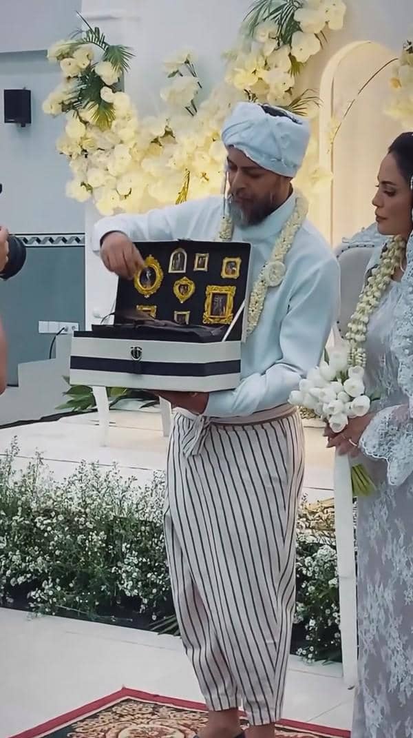 Seorang pria mengenakan pakaian adat berwarna putih dan sorban sedang menunjukkan kotak berisi perhiasan emas pada acara pernikahan.