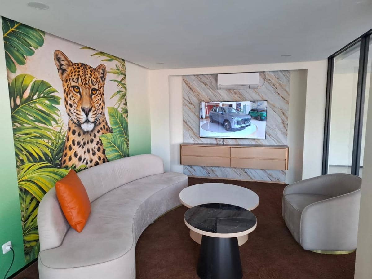 Ruang tunggu premium dengan sofa melengkung abu-abu, bantal oranye, mural macan tutul, dan televisi menampilkan mobil di dinding marmer.