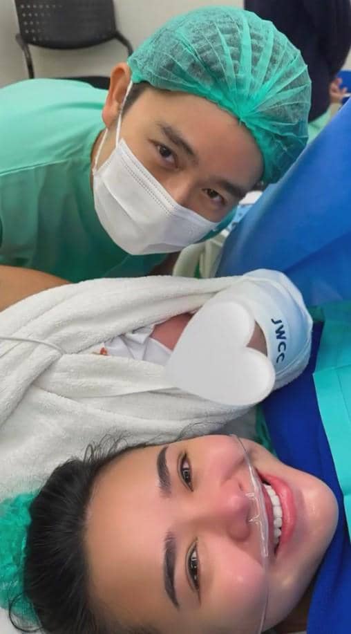 Seorang perempuan tersenyum di ranjang rumah sakit bersama bayi yang baru lahir dan seorang pria mengenakan pakaian medis serta masker.