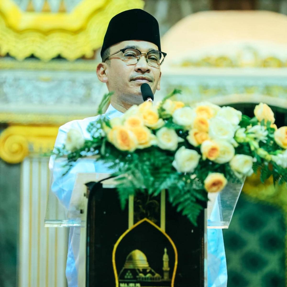 Seorang pria mengenakan peci hitam dan kemeja putih berbicara di podium berhias bunga mawar kuning dan putih di dalam ruangan bernuansa islami.
