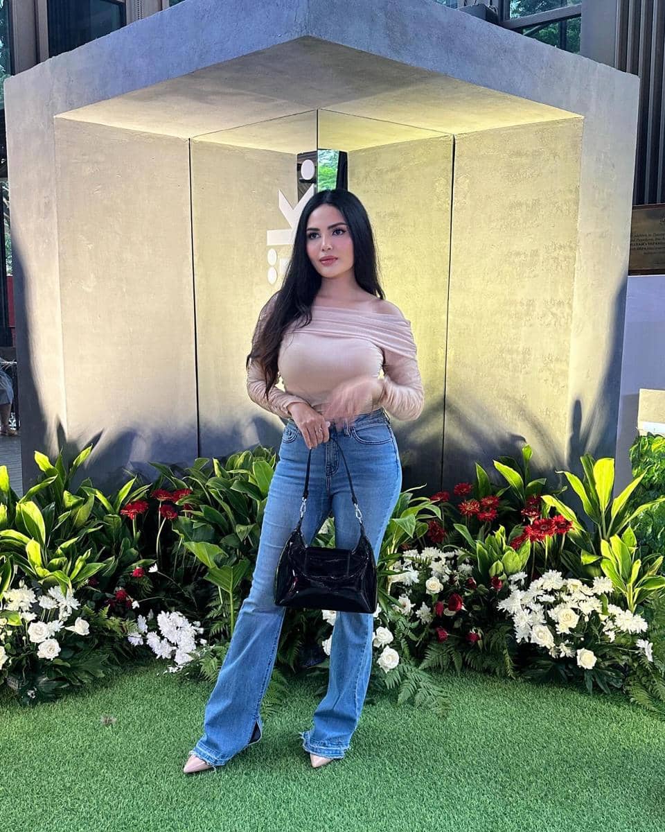 Seorang wanita berpose di depan instalasi beton dengan pencahayaan lembut, mengenakan atasan krem, celana jeans biru, dan tas hitam.