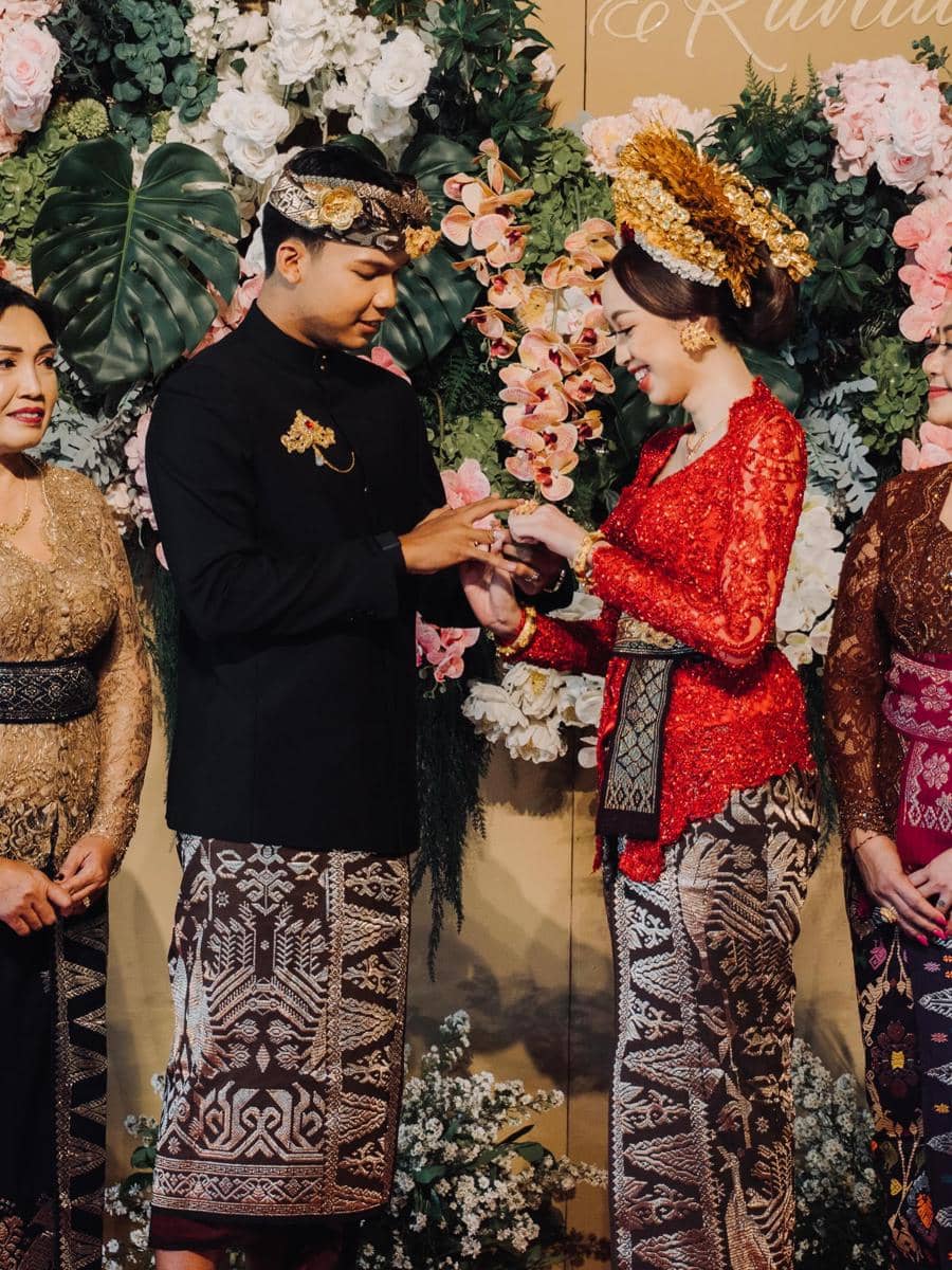 Pasangan mengenakan busana adat Jawa melakukan prosesi tukar cincin dalam acara lamaran dengan latar bunga berwarna cerah.