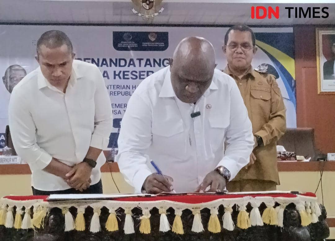 Seorang pejabat menandatangani dokumen di atas meja berhias kain tradisional, disaksikan dua pria lain di sebuah acara resmi di Kupang.