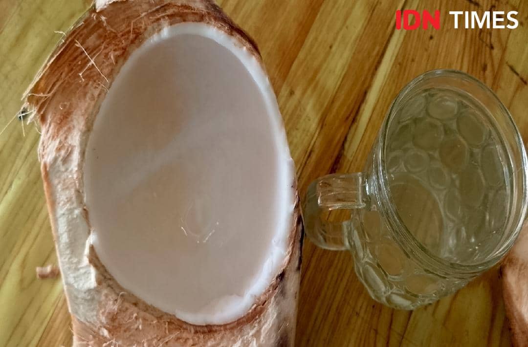 Setengah buah kelapa muda yang telah dibuka dengan gelas kaca berisi air kelapa di atas meja kayu.