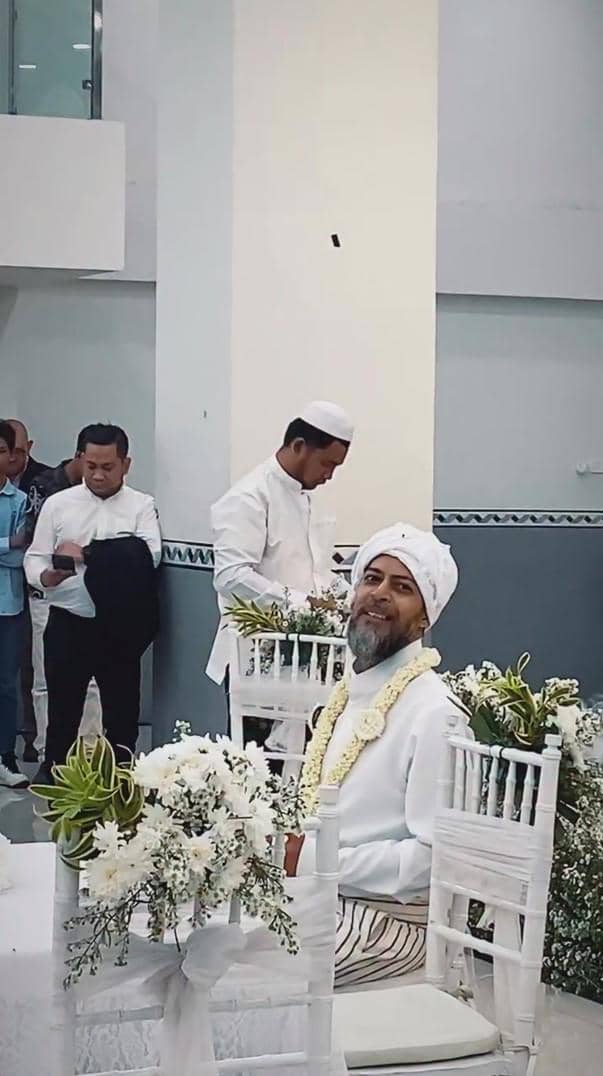 Seorang pria mengenakan pakaian serba putih dan sorban duduk di kursi berhias bunga putih dalam acara pernikahan bernuansa islami.