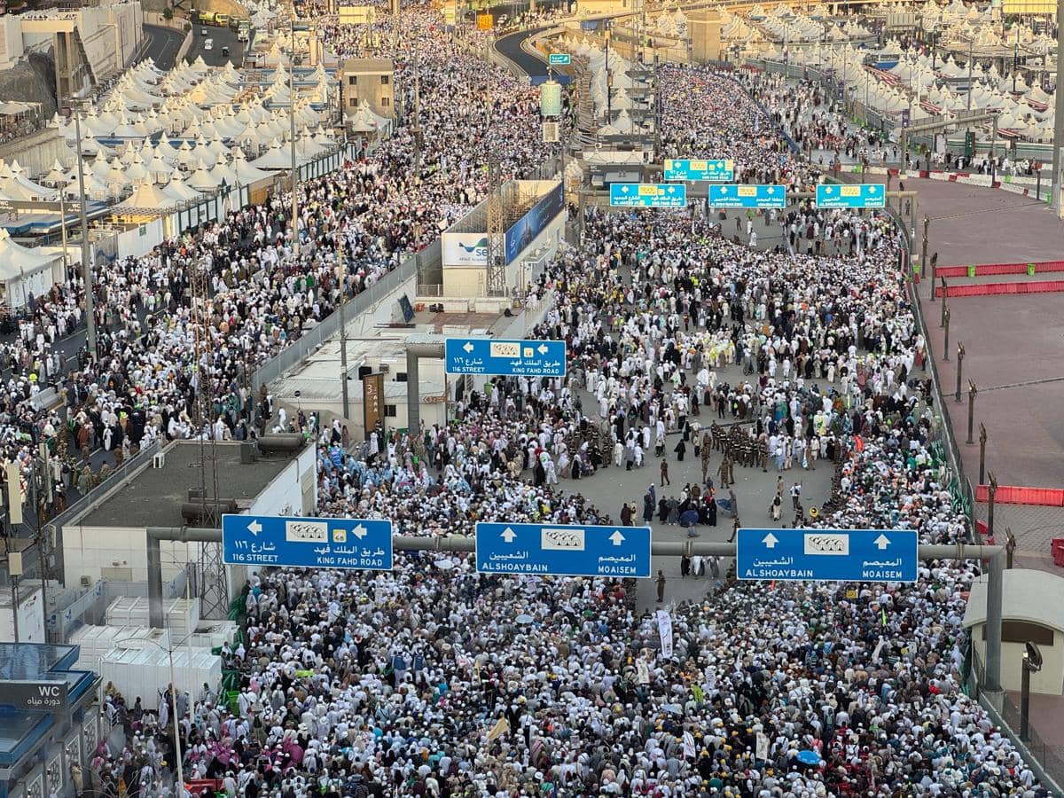 Ribuan jamaah haji berjalan padat dari area Jamarat menuju tenda-tenda di Mina dengan latar jalan dan papan penunjuk arah berbahasa Arab.