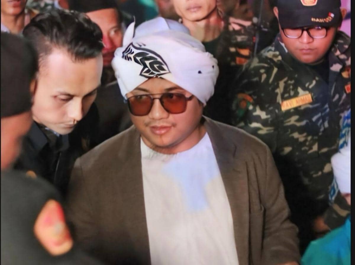 Seorang pria mengenakan sorban putih dan kacamata cokelat berjalan di tengah kerumunan bersama beberapa orang berseragam.