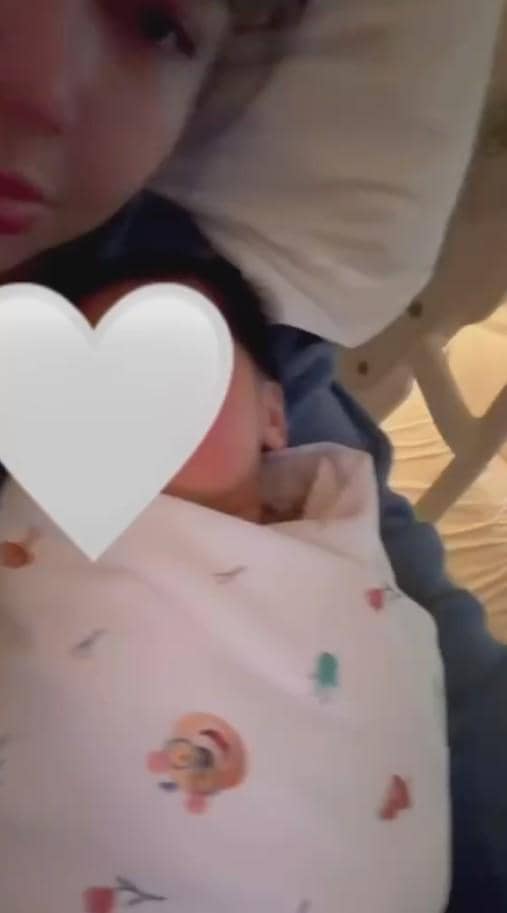 Seorang perempuan berbaring di tempat tidur bersama bayi yang dibedong selimut bermotif, wajah bayi ditutupi stiker berbentuk hati.