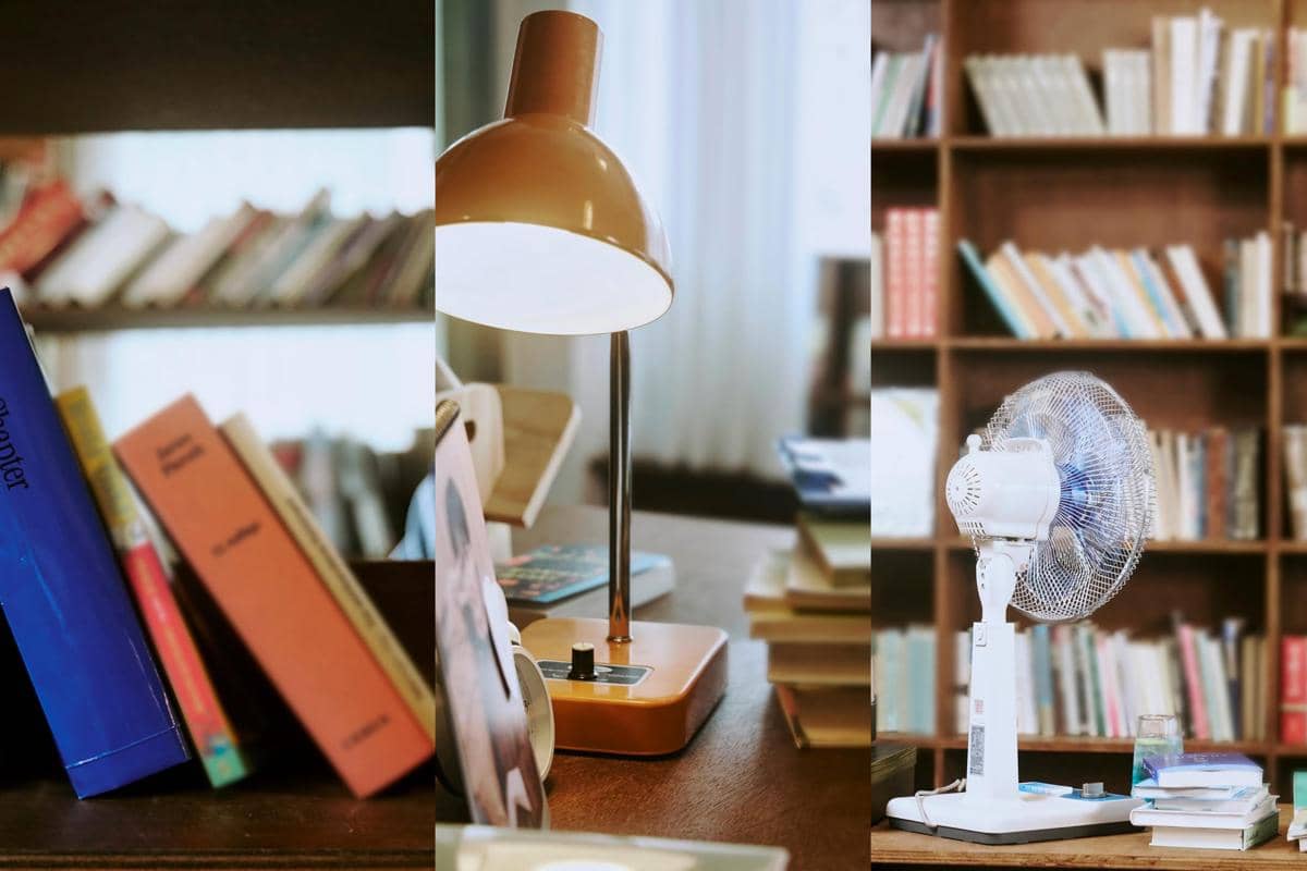Rak buku kayu dengan berbagai buku, meja berisi tumpukan buku, lampu meja berwarna oranye, dan kipas angin kecil di ruangan baca.