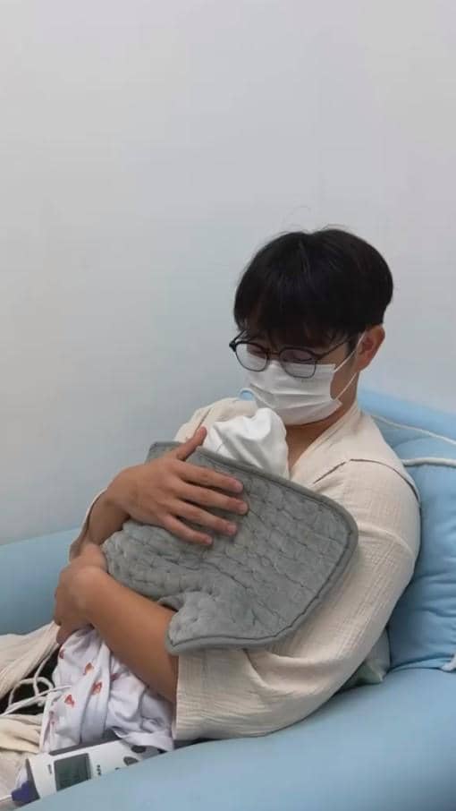 Seorang pria mengenakan masker dan kacamata duduk di sofa biru sambil menggendong bayi yang dibungkus selimut abu-abu di ruang bersih.