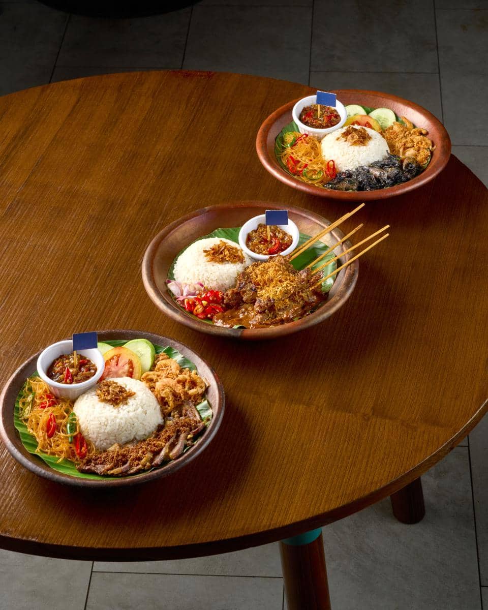 Tiga piring berisi nasi putih dengan lauk khas Jawa Timur, sambal, dan sayuran disajikan di atas meja kayu bundar.