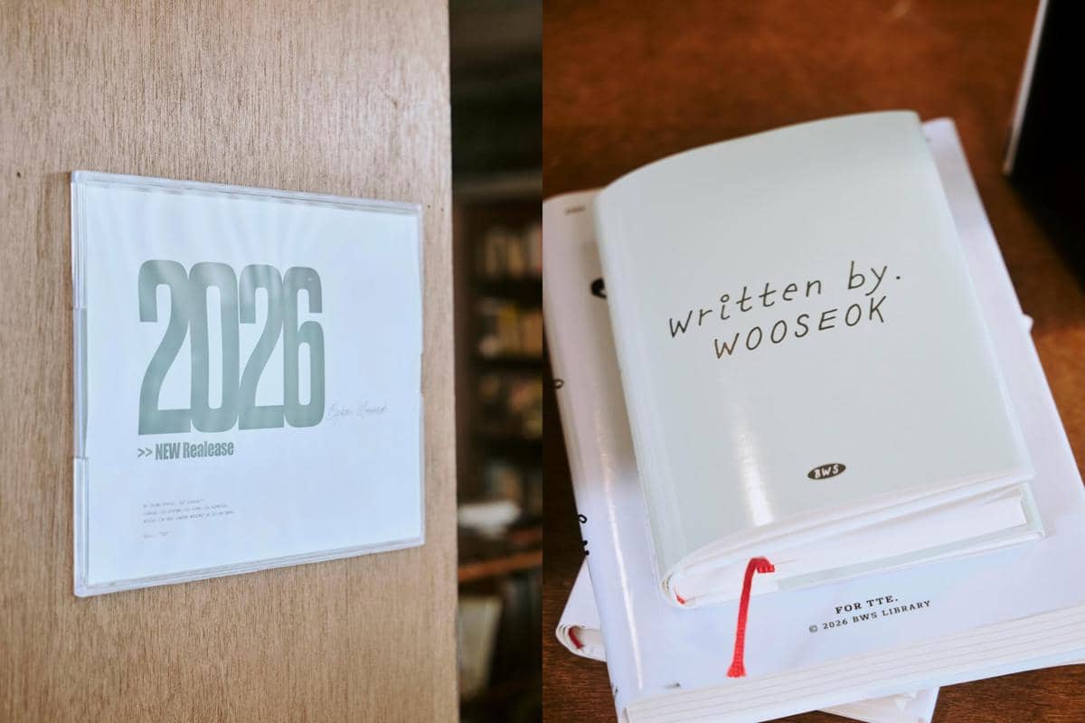 Sampul buku bertuliskan 'written by. WOOSEOK' di atas meja dan poster bertuliskan '2026 NEW Release' menempel di dinding kayu.
