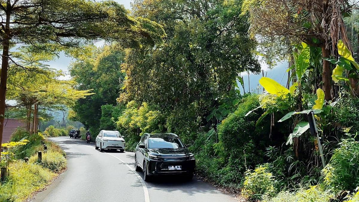 SUV terbaru DFSK berwarna hitam sedang menjalani uji jalan di jalan pedesaan Indonesia yang dikelilingi pepohonan hijau.