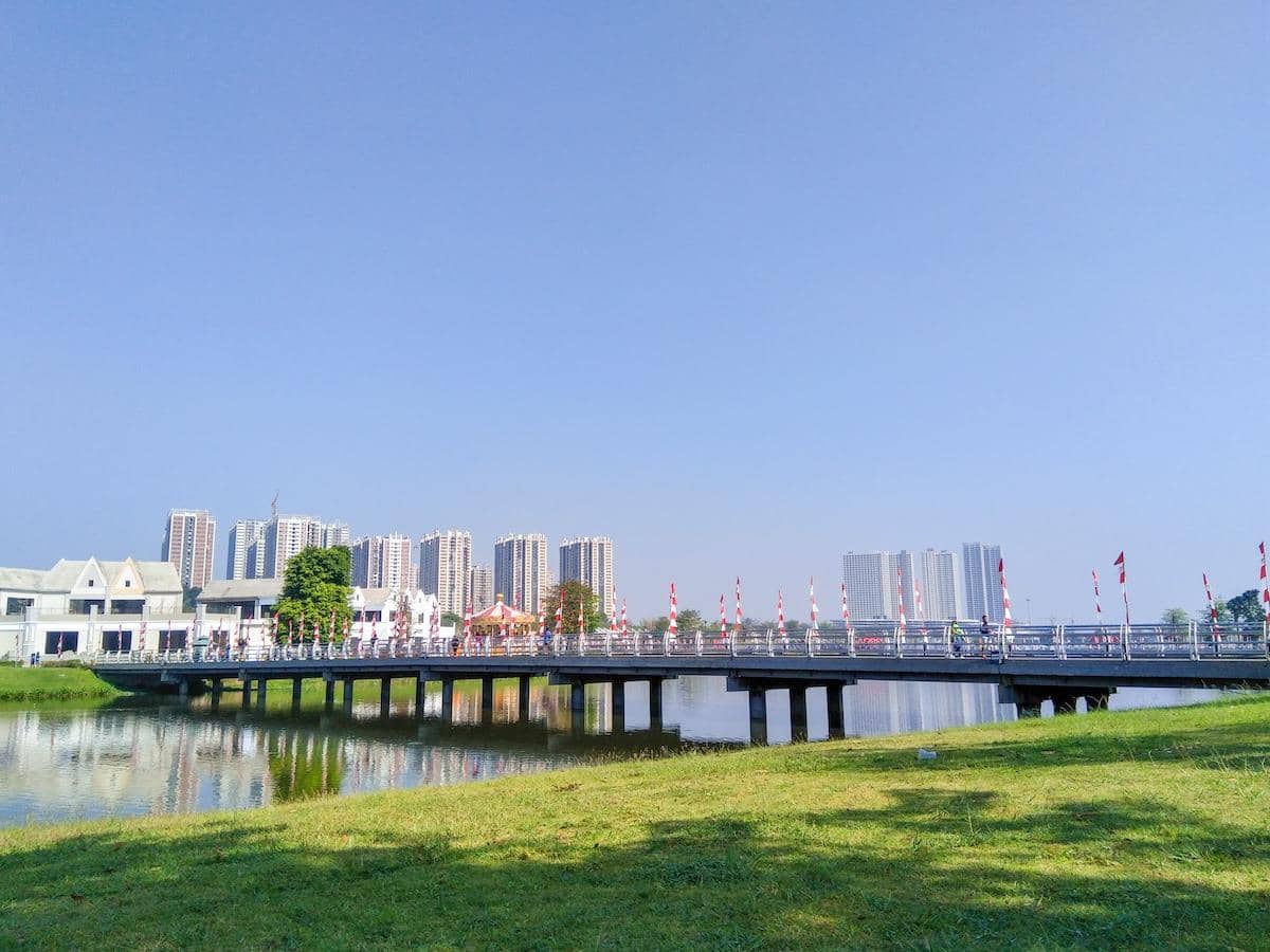 Pemandangan kawasan Cikarang dengan jembatan di atas danau, gedung tinggi di latar belakang, dan langit cerah berwarna biru.