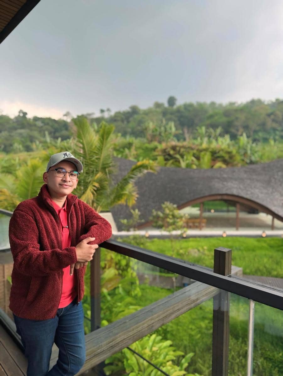 Seorang pria mengenakan jaket merah dan topi abu-abu berdiri di balkon dengan latar belakang pemandangan hijau dan bangunan beratap unik.