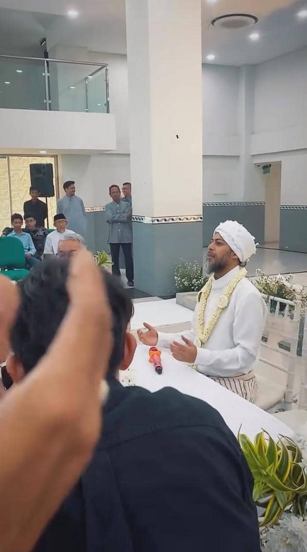 Seorang pria mengenakan pakaian putih dan sorban duduk di meja dengan mikrofon oranye di depannya dalam acara pernikahan di ruangan modern.